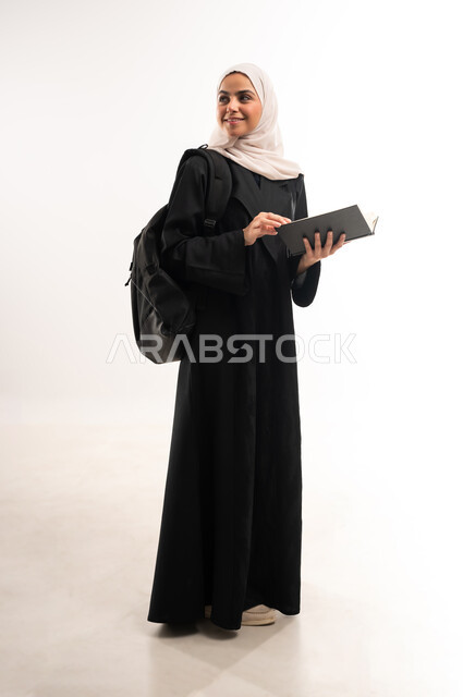 الاهتمام بتصفح الكتب المفيدة، التعليم الجامعي في المملكة, مراجعة الدروس اليومية, مفهوم الاجتهاد والتفوق, بورتريه لطالبة جامعية عربية خليجية سعودية محجبة ترتدي العباءة تحمل حقيبة على ظهرها، دراسة المحاضرات وتحضير البحوث العلمية, الطول الكامل للجسم، خلفية بيضاء