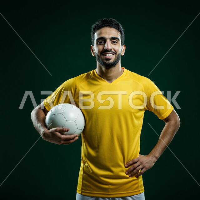 التحضير والتدريب و التجهز لبدء المباراة، رياضات شبابية وأنشطة ترفيهية، بورتريه مقرب للاعب عربي خليجي سعودي مبتسم يرتدي الزي الرياضي يحمل بيده كرة القدم ينظر الى الكاميرا بتعابير السرور والسعادة، خلفية خضراء