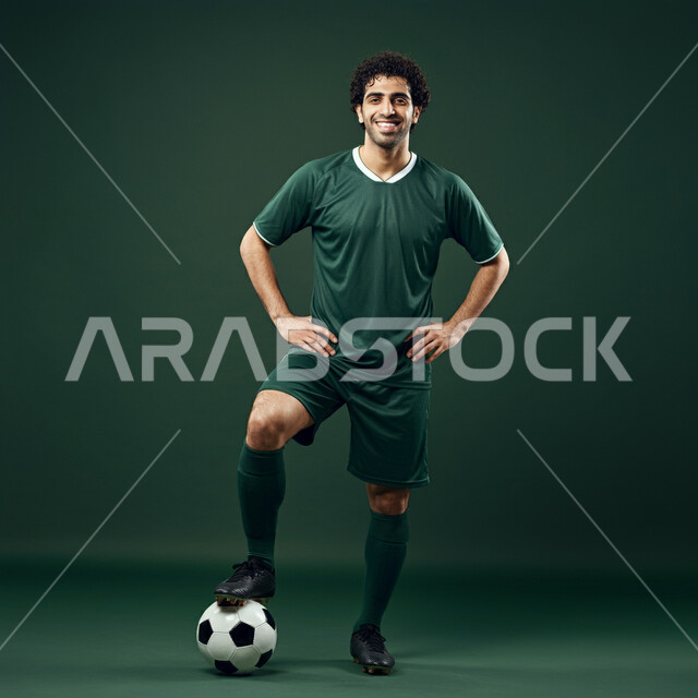 رياضات شبابية وأنشطة ترفيهية، بورتريه للاعب عربي خليجي سعودي مبتسم يرتدي الزي الرياضي يمسك الكرة بقدمه ينظر الى الكاميرا بتعابير السرور والسعادة، التحضير والتدريب و التجهز لبدء مباراة كرة القدم، صورة الجسم بالطول الكامل، خلفية خضراء