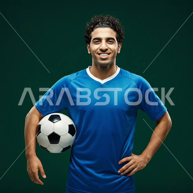 التدريب و التجهز والتحضير لبدء المباراة، رياضات شبابية وأنشطة ترفيهية، بورتريه مقرب للاعب عربي خليجي سعودي مبتسم يرتدي الزي الرياضي يحمل بيده كرة القدم ينظر الى الكاميرا بتعابير السرور والسعادة، خلفية خضراء