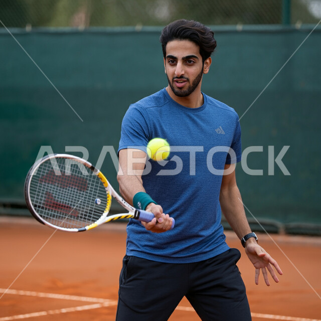 تعابير وايماءات تدل على الحماس والاندماج، شاب عربي خليجي سعودي يرتدي الملابس الرياضية يحمل بيده المضرب في الملعب باحتراف وإتقان، ممارسة لعبة كرة التنس، تدريبات وتمرينات رياضة المضرب، رياضات شبابية وأنشطة ترفيهية