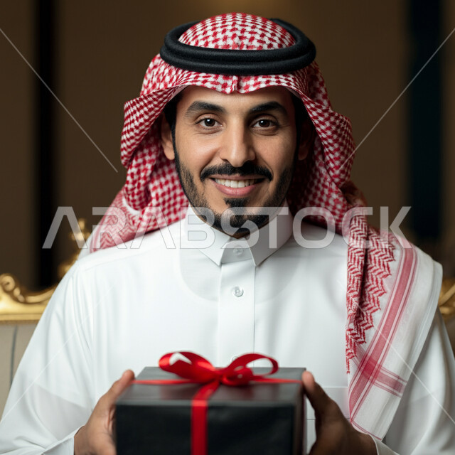  الأعياد والمناسبات السعيدة، النظر الى الكاميرا بتعابير السرور  والفرح، الاحتفال وتقديم الهدايا، صورة مقربة لرجل عربي سعودي مبتسم خليجي يرتدي الثوب التقليدي و الشماغ يحمل بيده علبة هدية مغلفة