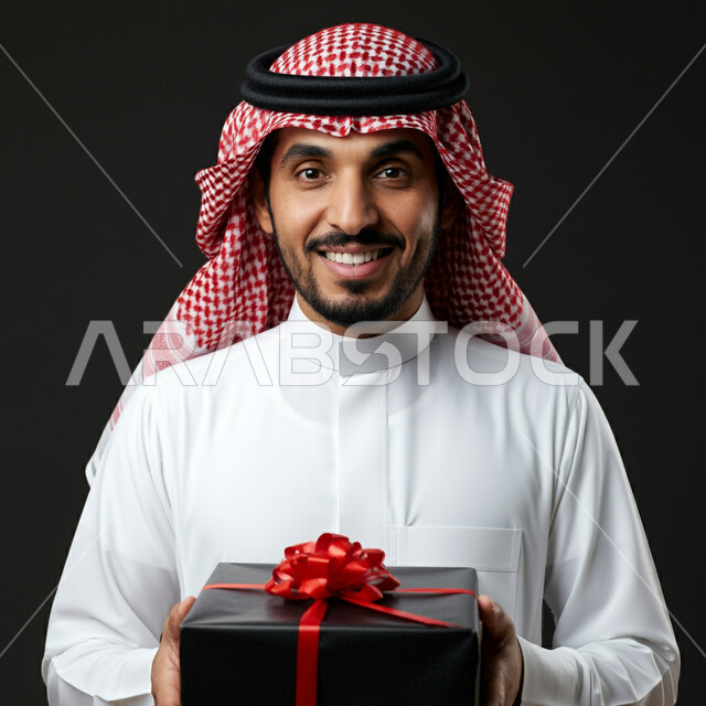 الاحتفال وتقديم الهدايا، الأعياد والمناسبات السعيدة، النظر الى الكاميرا بتعابير السرور  والفرح، بورتريه مقرب لرجل عربي سعودي مبتسم خليجي يرتدي الثوب التقليدي و الشماغ يحمل بيده علبة هدية مغلفة، خلفية رمادية
