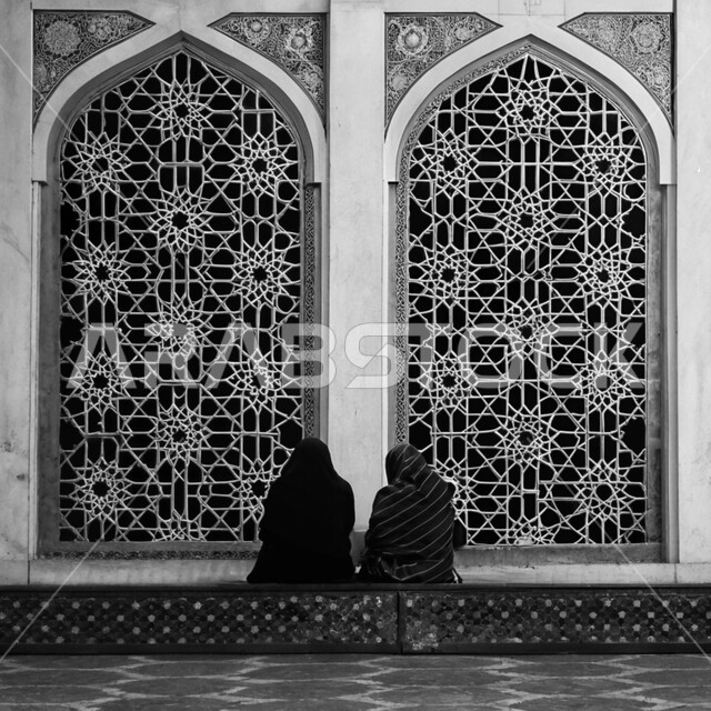 Worship and getting closer to God in the holy month of Ramadan, engravings and decorations on interior walls, concept of good company, black and white photo from the back of two women sitting inside a mosque in Saudi Arabia, Islamic architectural art