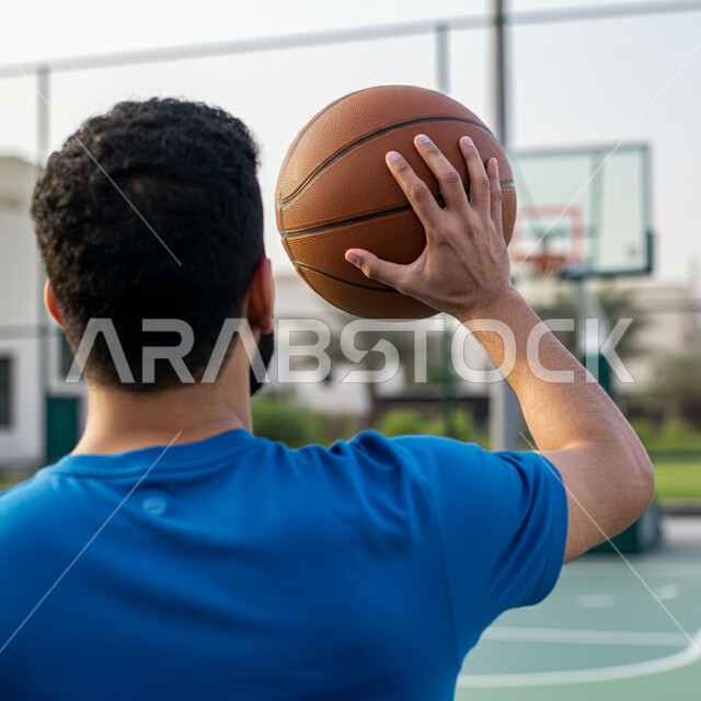 التجهز والتدريب والتحضير لبدء المباراة، مفهوم حب الرياضة، نمط الحياة الصحية، رياضات شبابية وأنشطة ترفيهية، صورة مقربة من الخلف للاعب عربي خليجي سعودي مبتسم يرتدي الزي الرياضي يحمل بيده كرة السلة 
