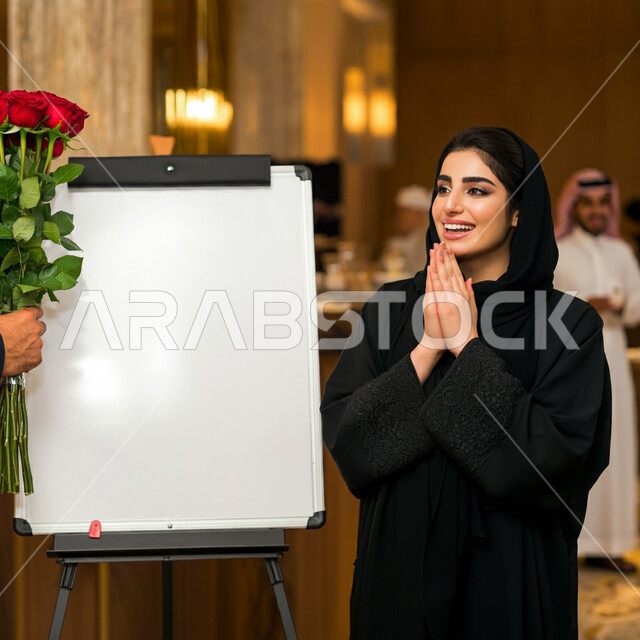  مفهوم الأنوثة والنعومة، الاهتمام بالجمال والأناقة والمظهر الخارجي، امرأة عربية خليجية سعودية ترتدي الحجاب والعباءة تقف امام موك ابيض مستطيل فارغ تحمل بيدها وردة حمراء جورية بايماءات الحب والفرح والسعادة، تقديم الزهور في الأعياد والمناسبات السعيدة