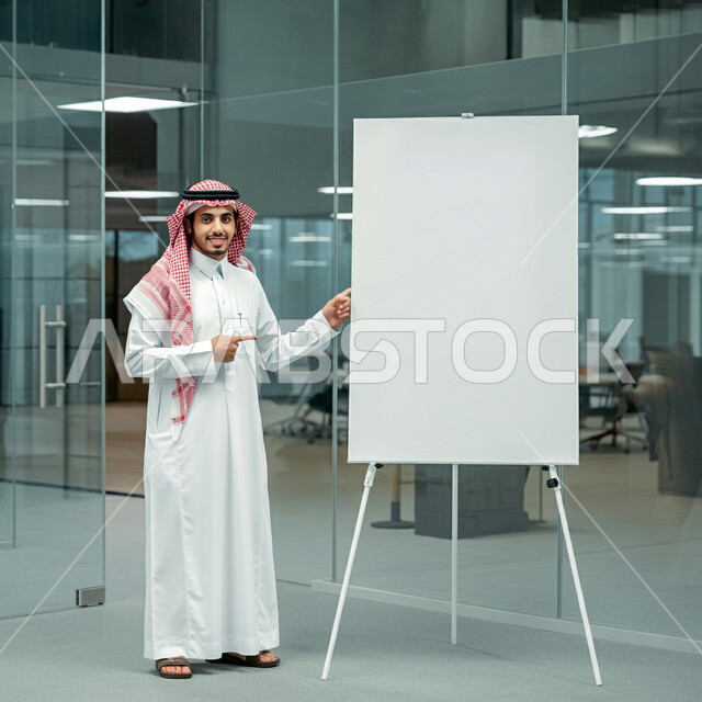 موك أب أبيض مستطيل، تقديم مقترحات المشاريع، رجل عربي خليجي سعودي يرتدي الثوب التقليدي والشماغ يشير باصبع السبابة الى لوح فارغ، عرض الأفكار الإبداعية، بيئة العمل المريحة في شركات السعودية، مناقشة الخطط المستقبلية، صورة لكامل الجسم