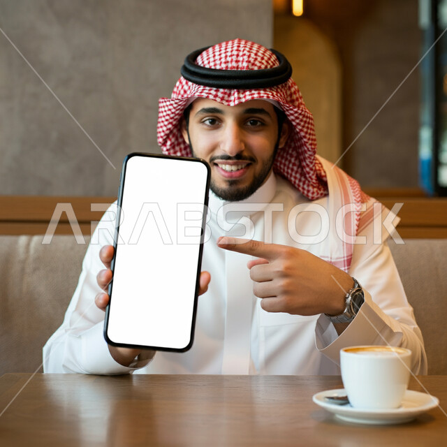 تناول المشروبات الساخنة، قضاء وقت ممتع في الخارج، موك أب أبيض مستطيل، صورة مقربة لرجل عربي خليجي سعودي يرتدي الثوب التقليدي والشماغ يجلس في مقهى يشير باصبع السبابة الى الهاتف المحمول، لوح اعلاني فارغ، النظر الى الكاميرا بايماءات السرور والسعادة
