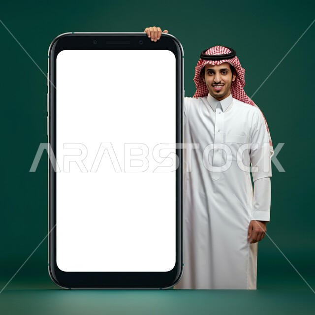 موك اب ابيض مستطيل، لوحة اعلانية فارغة، بورتريه لرجل عربي خليجي سعودي يرتدي الثوب التقليدي والشماغ  يقف بجانب مجسم لهاتف محمول بشاشة بيضاء ينظر الى الكاميرا بايماءات السرور والسعادة، خلفية خضراء
