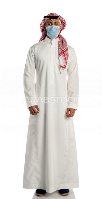 بورتريه لرجل عربي خليجي سعودي ينظر إلى الكاميرا، مرتدي القناع الطبي الوقائي للوقاية من فايروس كورونا كوفيد-19 ، خلفية بيضاء لرجل سعودي بالزي التقليدي السعودي مع شماغ وعقال ، إتباع إجراءات الوقاية من الفايروسات ، كامل الجسم ، صورة بالطول الكامل