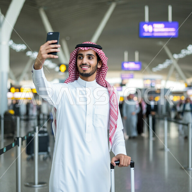 الوقوف في قاعة انتظار المطار، التقاط صور السيلفي التذكارية، قضاء أوقات ممتعه في العطلة الصيفيه، الاستعداد للسفر والرحلات السياحيه، وسائل النقل والمواصلات الجوية، رجل عربي خليجي سعودي يرتدي الثوب التقليدي والشماغ يمسك بحقيبة السفر يحمل بيده الهاتف المحمول