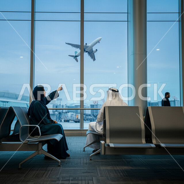 مشاهدة اقلاع الطائرات، صورة من الجانب لسيدة عربية خليجية سعودية تجلس في قاعة الانتظار في المطار تشير باصبع السبابة الى الطائرة، رحلة ترفيهية سياحية، توفير الخدمات المتطورة لسهولة التنقل بين البلدان، شركات وخطوط الطيران السعودي