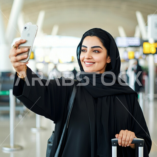 التقاط صور السيلفي التذكارية، قضاء أوقات ممتعه في العطلة الصيفيه، الاستعداد للسفر والرحلات السياحيه، وسائل النقل والمواصلات الجوية، صورة مقربة لشابة عربية خليجية سعودية محجبة تقف في قاعة انتظار المطار تمسك بحقيبة السفر تحمل بيدها الهاتف المحمول