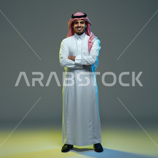 The concept of elegance and masculinity, standing straight and confident, portrait of a handsome young Saudi Arabian Gulf man smiling wearing a shemagh and traditional dress, looking at the camera with crossed hand gestures, taking care of one's appearance, full-length body image, colored background