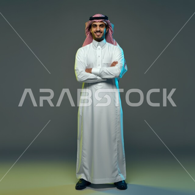 The concept of elegance and masculinity, standing straight and confident, portrait of a handsome young Saudi Arabian Gulf man smiling wearing a shemagh and traditional dress, looking at the camera with crossed hand gestures, taking care of one's appearance, full-length body image, colored background