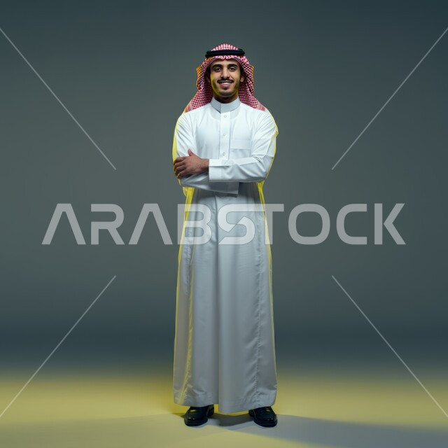 النظر إلى الكاميرا بإيماءات أيدي متقاطعه، مفهوم الاناقه والرجولة ،الوقوف باستقامة وثقه بالنفس، بورتريه لشاب وسيم عربي خليجي سعودي مبتسم يرتدي الشماغ والثوب التقليدي، الاهتمام بالمظهر الخارجي، صورة الجسم بالطول الكامل، خلفية ملونة