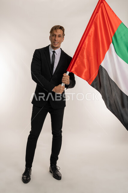 التعرف على ثقافات وتاريخ الامارات، بورتريه لرجل اعمال اجنبي مبتسم يرتدي البدلة الرسمية يحمل العلم للاحتفال باليوم الوطني الامارتي2ديسمبر ، النظر الى الكاميرا بتعابير الفخر والسرور، ذكرى تأسيس الاتحاد1971 ، صورة الجسم بالكامل، خلفية بيضاء