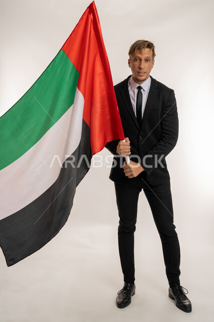 التعرف على ثقافات وتاريخ الامارات، بورتريه لرجل اعمال اجنبي مبتسم يرتدي البدلة الرسمية يحمل العلم للاحتفال باليوم الوطني الامارتي2ديسمبر ، النظر الى الكاميرا بتعابير الفخر والسرور، ذكرى تأسيس الاتحاد1971 ، صورة الجسم بالكامل، خلفية بيضاء