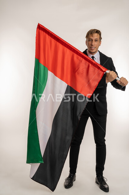 Getting to know the culture and history of the Emirates, portrait of a smiling foreign businessman in a formal suit holding a flag to celebrate the UAE National Day, December 2, looking at the camera with expressions of pride and joy, anniversary of the founding of the Union 1971, full body photo, white background