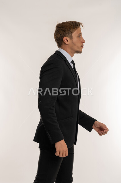 النظر الى شيء ما بتعابير الحزم والجدية، ايماءات المشي و الذهاب إلى مكان معين، مفهوم الاناقة والرجولة والاهتمام بالمظهر الخارجي، بورتريه من الجانب لرجل اعمال اجنبي يرتدي البدلة الرسمية يقوم بالتقدم الى الامام بخطوات ثابتة، خلفية بيضاء