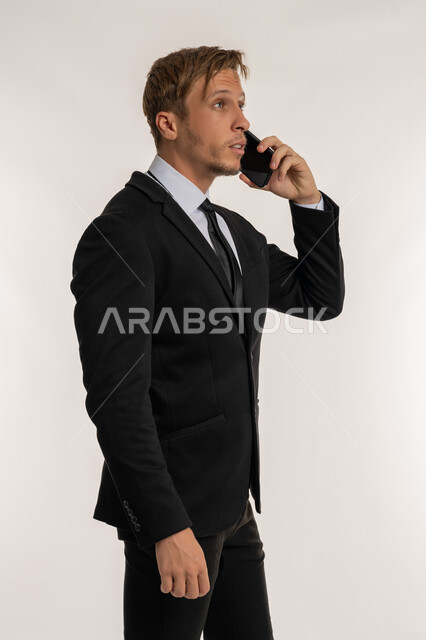 ايماءات السعادة والسرور لسماع الاخبار المفرحة، بورتريه من الجانب لرجل اعمال اجنبي مبتسم يرتدي البدلة الرسمية يقوم باجراء مكالمة هاتفية عبر الهاتف المحمول، استخدام التقنيات والاجهزة الحديثة، التواصل مع الاهل والاصدقاء، خلفية بيضاء