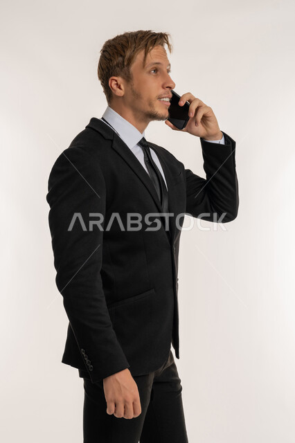 ايماءات السعادة والسرور لسماع الاخبار المفرحة، بورتريه من الجانب لرجل اعمال اجنبي مبتسم يرتدي البدلة الرسمية يقوم باجراء مكالمة هاتفية عبر الهاتف المحمول، استخدام التقنيات والاجهزة الحديثة، التواصل مع الاهل والاصدقاء، خلفية بيضاء