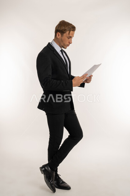 التدقيق في قراءة الملاحظات، اعداد التقارير الخاصة بالعمل، تعابير الفتكير والتركيز، بورتريه من الجانب لرجل اعمال اجنبي يرتدي البدلة الرسمية يحمل بيده مجموعة من الوثائق والاوراق البيضاء، مراجعة ومتابعة الخطط والمشاريع  بإيماءات السعادة، صورة الجسم بالكامل، خلفية بيضاء