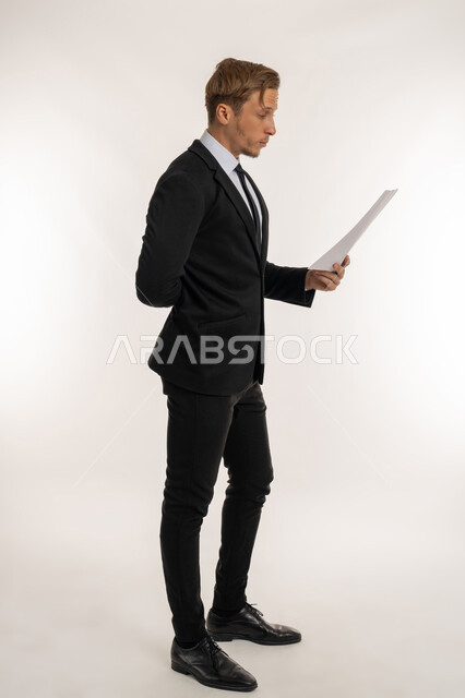 التدقيق في قراءة الملاحظات، اعداد التقارير الخاصة بالعمل، تعابير الفتكير والتركيز، بورتريه من الجانب لرجل اعمال اجنبي يرتدي البدلة الرسمية يحمل بيده مجموعة من الوثائق والاوراق البيضاء، مراجعة ومتابعة الخطط والمشاريع  بإيماءات السعادة، صورة الجسم بالكامل، خلفية بيضاء