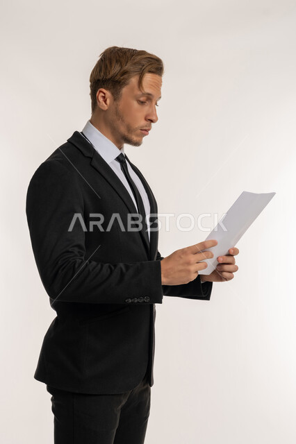 تعابير الفتكير والتركيز، التدقيق في قراءة الملاحظات، اعداد التقارير الخاصة بالعمل، بورتريه من الجانب لرجل اعمال اجنبي يرتدي البدلة الرسمية يحمل بيده مجموعة من الوثائق والاوراق البيضاء، مراجعة ومتابعة الخطط والمشاريع  بإيماءات السعادة والسرور، خلفية بيضاء