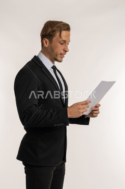 تعابير الفتكير والتركيز، التدقيق في قراءة الملاحظات، اعداد التقارير الخاصة بالعمل، بورتريه من الجانب لرجل اعمال اجنبي يرتدي البدلة الرسمية يحمل بيده مجموعة من الوثائق والاوراق البيضاء، مراجعة ومتابعة الخطط والمشاريع  بإيماءات السعادة والسرور، خلفية بيضاء