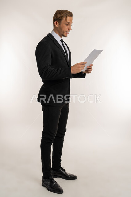 تعابير الفتكير والتركيز، التدقيق في قراءة الملاحظات، اعداد التقارير الخاصة بالعمل، بورتريه من الجانب لشاب اجنبي يرتدي الثوب الرسمي يحمل بيده مجموعة من الوثائق والاوراق البيضاء، مراجعة ومتابعة الخطط والمشاريع بايماءات السعادة، صورة الجسم بالكامل، خلفية بيضاء