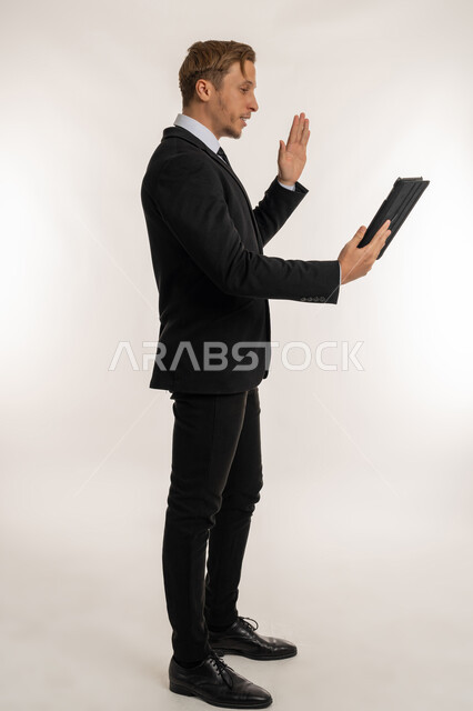 القاء التحية والترحيب والسلام، بورتريه من الجانب لرجل اعمال اجنبي يرتدي البدلة الرسمية يقوم بإجراء مكالمة فيديو باستخدام جهاز التابلت، التواصل مع العملاء والاهل والاصدقاء، دمج التقنية والتكنولوجيا في الحياة اليومية، عقد اجتماع عن بعد، صورة الجسم بالكامل، خلفية بيضاء