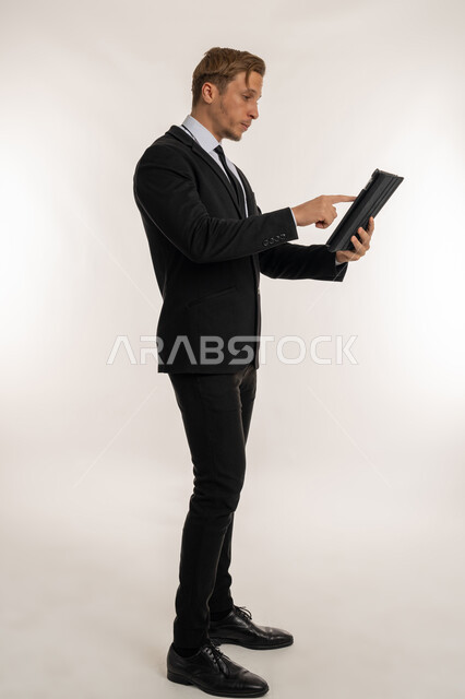 Browsing the internet and completing tasks remotely, following the latest social media news, holding a business meeting, integrating technology and technology into daily life, side portrait of foreign businessman in formal suit using tablet with expressions of concentration, full body image, white background