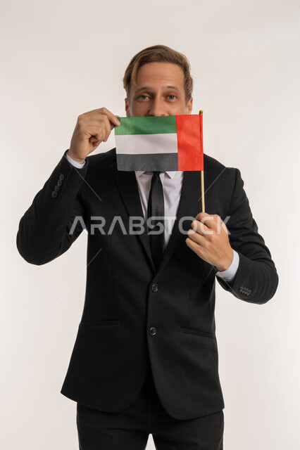 الاحتفال بالاعياد الوطنية للشعوب، اليوم الوطني الاماراتي2 ديسمبر، بورتريه لرجل اجنبي يرتدي البدلة الرسمية يقوم بوضع علم الامارات أمام وجهه، الوقوف باستقامة والنظر إلى الكاميرا بإيماءات الفخر والسرور، إحياء ذكرى استقلال البلاد، خلفية بيضاء