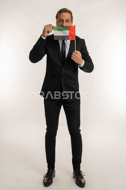 إحياء ذكرى استقلال البلاد، الاحتفال بالاعياد الوطنية للشعوب، اليوم الوطني الاماراتي2 ديسمبر، بورتريه لرجل اجنبي يرتدي البدلة الرسمية يقوم بوضع علم الامارات أمام وجهه، الوقوف باستقامة والنظر إلى الكاميرا بإيماءات الفخر والسرور، صورة الجسم بالكامل، خلفية بيضاء