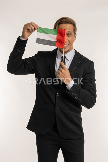إحياء ذكرى استقلال البلاد، الاحتفال بالاعياد الوطنية للشعوب، اليوم الوطني الاماراتي2 ديسمبر، بورتريه لرجل اجنبي يرتدي البدلة الرسمية يقوم بوضع علم الامارات أمام وجهه، الوقوف باستقامة والنظر إلى الكاميرا بإيماءات الفخر والسرور، خلفية بيضاء