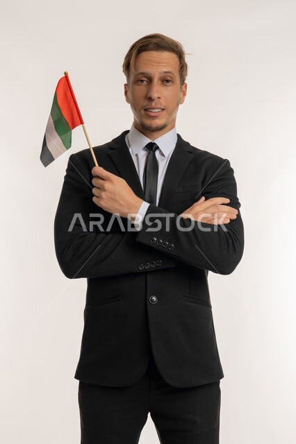 التعرف على ثقافات وشعب الامارات، الوقوف بإيماءات ايدي متقاطعة، إحياء ذكرى استقلال البلاد، الاحتفال باليوم الوطني الاماراتي2ديسمبر، بورتريه لرجل اجنبي يرتدي البدلة الرسمية يمسك بيده العلم الاماراتي، النظر إلى الكاميرا بتعابير الفخر والسرور، خلفية بيضاء