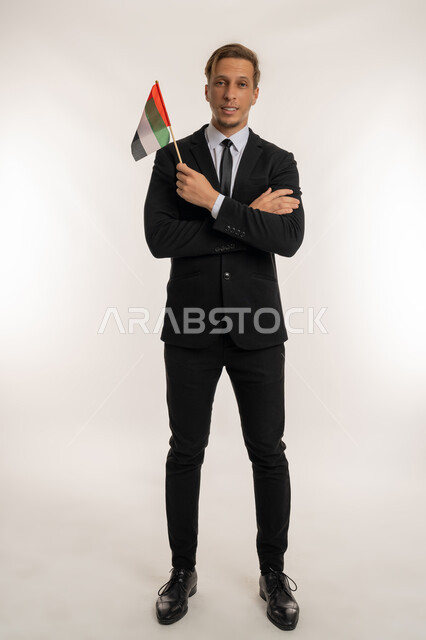 الوقوف بإيماءات ايدي متقاطعة، إحياء ذكرى استقلال البلاد، اليوم الوطني الاماراتي2 ديسمبر، بورتريه لرجل اجنبي يرتدي البدلة الرسمية يمسك بيده علم الامارات المتحدة، النظر إلى الكاميرا بتعابير الفخر، الاحتفال بالاعياد الوطنية للشعوب، صورة الجسم بالكامل، خلفية بيضاء