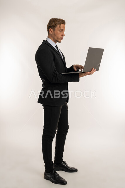 النقر والتفاعل مع لوحة المفاتيح، عقد إجتماع عمل عبر الإنترنت، بورتريه من الجانب لرجل اعمال اجنبي يرتدي البدلة الرسمية يقوم بالعمل على الحاسب المحمول, دمج الاجهزة الحديثة والتكنولوجيا في الحياة العملية، إدارة شؤون الأعمال وتنظيمها، صورة الجسم بالكامل، خلفية بيضاء