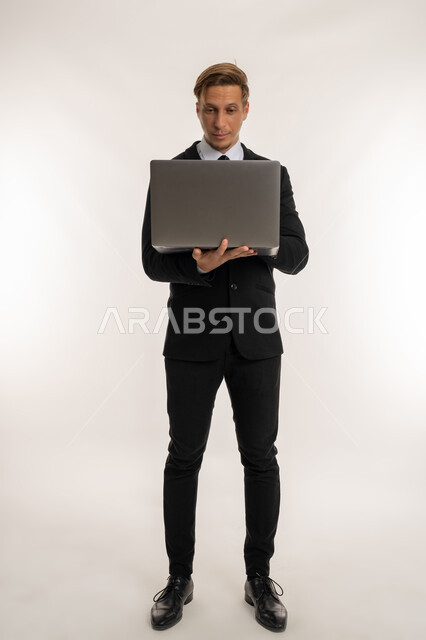 إدارة شؤون الأعمال وتنظيمها، استخدام التقنيات الحديثة المتطورة، عقد إجتماع عمل عبر الإنترنت، بورتريه لرجل اعمال اجنبي يرتدي البدلة الرسمية يقوم بالعمل على الحاسب المحمول, دمج التكنولوجيا في الحياة العملية، صورة الجسم بالكامل، خلفية بيضاء