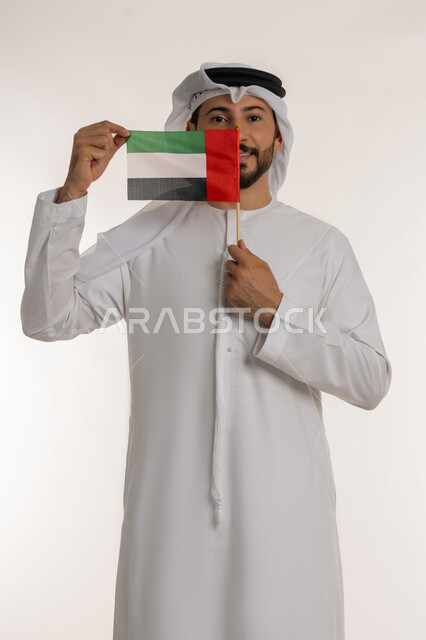 إحياء ذكرى استقلال البلاد1971، بورتريه لرجل عربي خليجي إماراتي يرتدي الكندورة والغترة يضع علم دولة الامارات العربية المتحدة امام وجهه، الاحتفال باليوم الوطني 2 ديسمبر، النظر إلى الكاميرا بإيماءات الاعتزاز والفخر، مفهوم حب الوطن والانتماء إليه،  خلفية بيضاء
