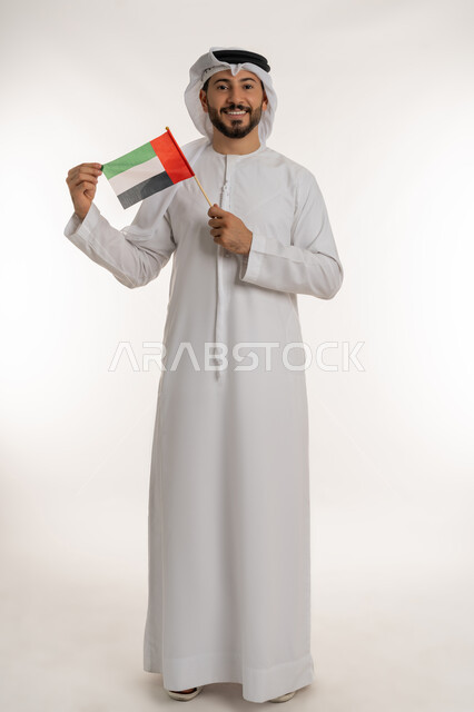 مفهوم حب الوطن والانتماء إليه، إحياء ذكرى استقلال البلاد1971، بورتريه لرجل عربي خليجي إماراتي يرتدي الكندورة والغترة يرفع علم الإمارات العربية المتحدة، الاحتفال باليوم الوطني 2 ديسمبر، النظر إلى الكاميرا بإيماءات الاعتزاز والفخر، صورة الجسم بالكامل، خلفية بيضاء
