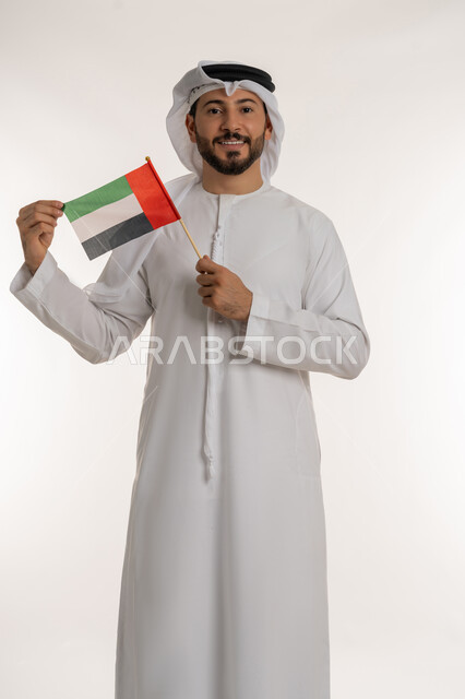 إحياء ذكرى استقلال البلاد1971، بورتريه لرجل عربي خليجي إماراتي يرتدي الكندورة والغترة يضع علم دولة الامارات العربية المتحدة امام وجهه، الاحتفال باليوم الوطني 2 ديسمبر، النظر إلى الكاميرا بإيماءات الاعتزاز والفخر، مفهوم حب الوطن والانتماء إليه،  خلفية بيضاء