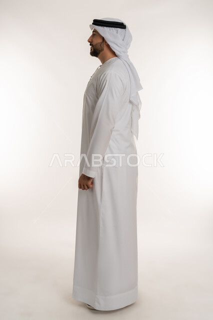 مفهوم الأناقة والرجولة والقوة, الاهتمام بالمظهر الخارجي، تعابير السرور والسعادة، الوقوف باستقامة والنظر الى مكان ما بايماءات الثقة بالنفس, بورتريه من الجانب لرجل عربي خليجي إماراتي يرتدي الكندورة الاماراتية والغترة ينظر الى شيء ما، صورة لكامل الجسم، خلفية بيضاء