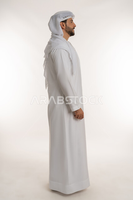 الاهتمام بالمظهر الخارجي، تعابير السرور والسعادة، الوقوف باستقامة والنظر الى مكان ما بايماءات الثقة بالنفس, مفهوم الأناقة والرجولة والقوة, بورتريه من الجانب لرجل عربي خليجي إماراتي يرتدي الكندورة الاماراتية والغترة ينظر الى شيء ما، صورة لكامل الجسم، خلفية بيضاء