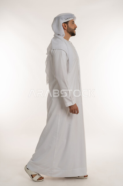 التقدم إلى الأمام بخطوات ثابتة، مفهوم الأناقة والرجولة والقوة, الاهتمام بالمظهر الخارجي، ايماءات المشي و الذهاب إلى مكان معين، بورتريه من الجانب لرجل عربي خليجي اماراتي يرتدي الكندورة والغترة ينظر إلى شيء ما بتعابير السرور والسعادة، صورة لكامل الجسم، خلفية بيضاء