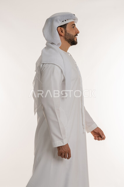 الاهتمام بالمظهر الخارجي، التقدم إلى الأمام بخطوات ثابتة، مفهوم الأناقة والرجولة والقوة, ايماءات المشي و الذهاب إلى مكان معين، بورتريه من الجانب لرجل عربي خليجي اماراتي يرتدي الكندورة والغترة ينظر إلى شيء ما بتعابير السرور والسعادة، خلفية بيضاء
