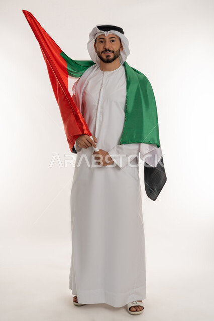 إحياء ذكرى استقلال البلاد1971، مفهوم حب الوطن والانتماء إليه، بورتريه لرجل عربي خليجي إماراتي يرتدي الكندورة والغترة يضع علم الإمارات العربية المتحدة على كتفيه، الاحتفال باليوم الوطني 2 ديسمبر، النظر إلى الكاميرا بإيماءات الاعتزاز، صورة لكامل الجسم، خلفية بيضاء