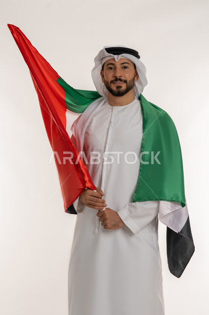 الاحتفال باليوم الوطني 2 ديسمبر، إحياء ذكرى استقلال البلاد1971، مفهوم حب الوطن والانتماء إليه، بورتريه لرجل عربي خليجي إماراتي يرتدي الكندورة والغترة يضع علم الإمارات العربية المتحدة على كتفيه، النظر إلى الكاميرا بإيماءات الاعتزاز، خلفية بيضاء