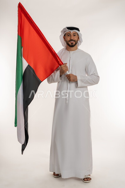 The concept of patriotism and belonging to the homeland, commemorating the country's independence in 1971, a portrait of an Emirati Gulf Arab man wearing a kandura and ghutra raising the flag of the United Arab Emirates, celebrating the National Day on December 2, looking at the camera with gestures of pride and honor, a full body photo, white background